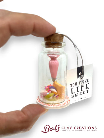 Miniature dessert in a bottle with a cork lid, held by a hand, on a white background with 'Bert's Clay Creations' branding.